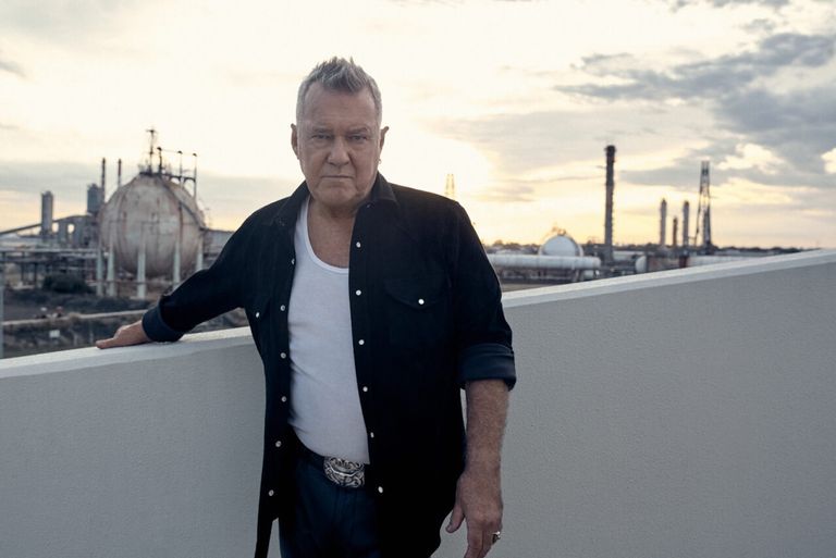 Australian rock musician Jimmy Barnes posing outdoors in an unbuttoned shirt and white singlet with an industrial plant in the background.